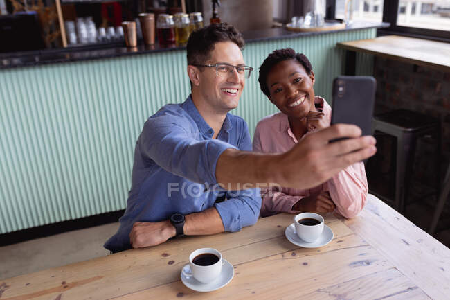 In der Mitte lächelt ein gemischtes Paar, während es ein Selfie beim Kaffeetrinken in einem Café macht. Paardatum und Beziehungskonzept — Stockfoto
