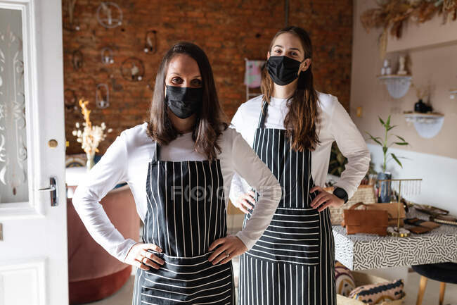 Kaukasische Geschäftsinhaberin und Kellnerin tragen Gesichtsmasken und Schürzen und blicken in die Kamera. kleines unabhängiges Café-Geschäft während der Coronavirus-Pandemie 19. — Stockfoto
