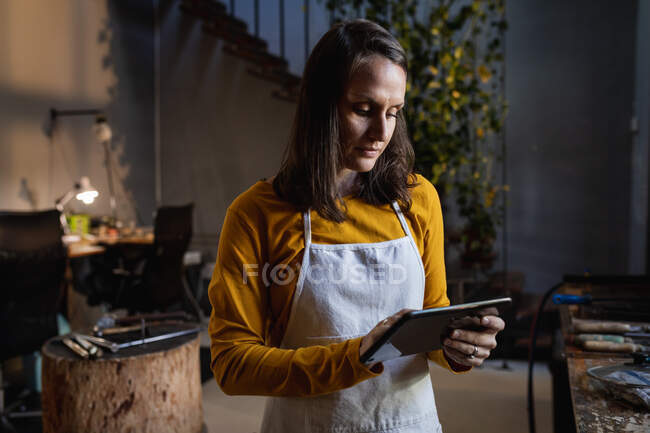 Focused caucasian female jeweller wearing apron, standing in workshop, using tablet. independent handmade craft business. — Stock Photo