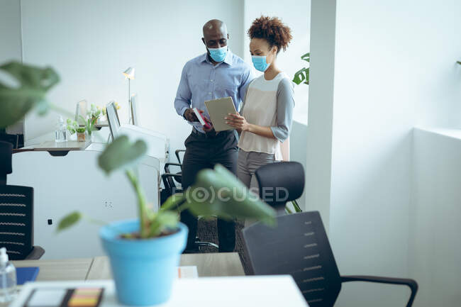 Two diverse business colleagues wearing face masks and using tablet. work at a modern office during covid 19 coronavirus pandemic. — Stock Photo