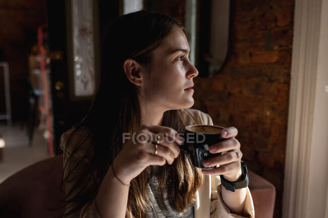 Cliente femenino caucásico sentado en la mesa, mirando fuera de la ventana, bebiendo café. pequeño negocio de café independiente. - foto de stock