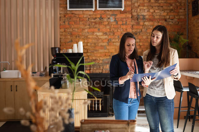 Due donne caucasiche in piedi all'interno del caffè, tenendo i menu e parlando. piccola impresa indipendente caffè. — Foto stock