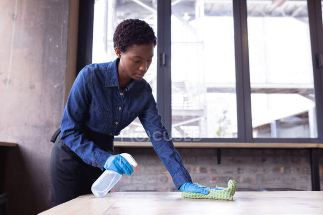 Femme afro-américaine nettoyant la table avec du désinfectant pulvérisateur et un chiffon au café. nettoyage et désinfection pour la prévention et le contrôle de l'épidémie de covide-19 — Photo de stock