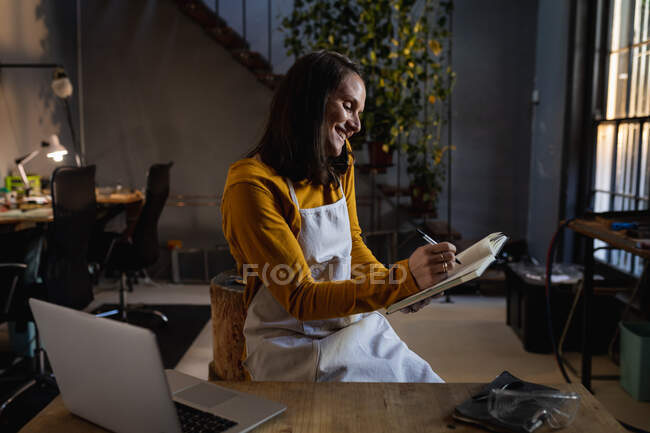 Улыбающаяся белая ювелирка в фартуке, сидящая за столом, используя смартфон, делая заметки. независимый ремесленный бизнес. — стоковое фото