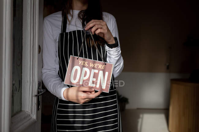 Sección media de la camarera con delantal a rayas, de pie en la puerta, sosteniendo el cartel abierto. pequeño negocio de café independiente. - foto de stock
