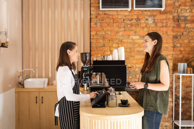 Dos mujeres caucásicas propietarias de negocios y cliente de pie en la encimera, hablando y sonriendo. pequeño negocio de café independiente. - foto de stock