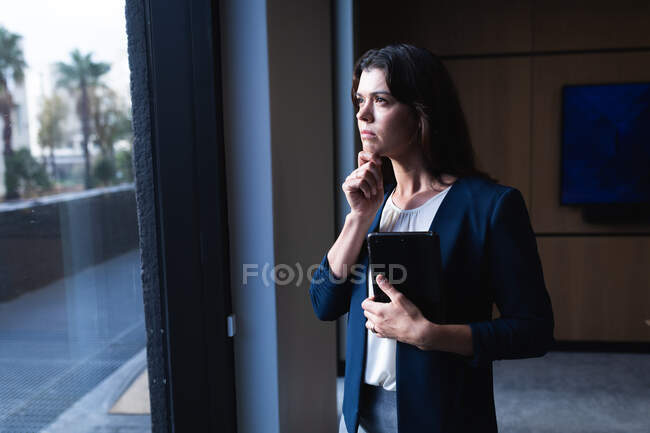 Nachdenkliche kaukasische Geschäftsfrau, die ein digitales Tablet in der Hand hält und aus dem Fenster auf das moderne Büro blickt. Geschäfts- und Bürokonzept — Stockfoto