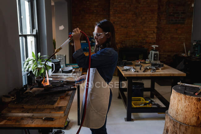Caucasian female jeweller wearing apron and glasses, using gas burner, melting metal for jewelry. independent handmade craft business. — Stock Photo