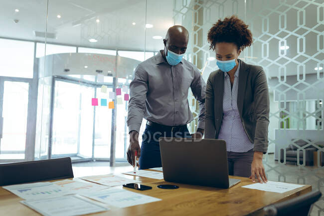 Дві різні чоловічі та жіночі бізнес-колеги носять маски для обличчя та використовують ноутбук. робота в сучасному офісі під час пандемії коронавірусу 19 . — стокове фото