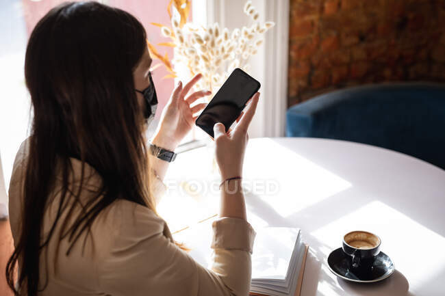 Caucasian female customer wearing face mask sitting at table, using smartphone. small independent cafe business during coronavirus covid 19 pandemic. — Stock Photo