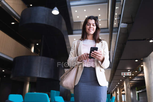 Donna d'affari caucasica sorridente durante l'utilizzo di smartphone mentre in piedi in ufficio moderno. concetto di business e ufficio — Foto stock
