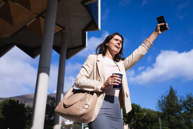 Donna d'affari caucasica con in mano una tazza di caffè e uno smartphone che chiama un taxi per strada. concetto di business e ufficio — Foto stock