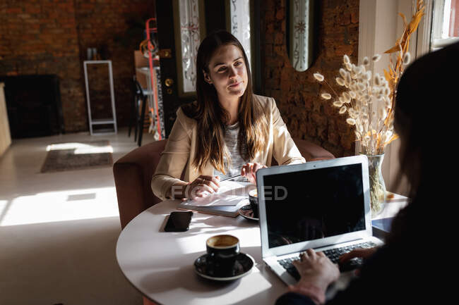 Zwei kaukasische Frauen sitzen bei Kaffee am Tisch, benutzen Laptop und unterhalten sich. kleines unabhängiges Café-Geschäft. — Stockfoto