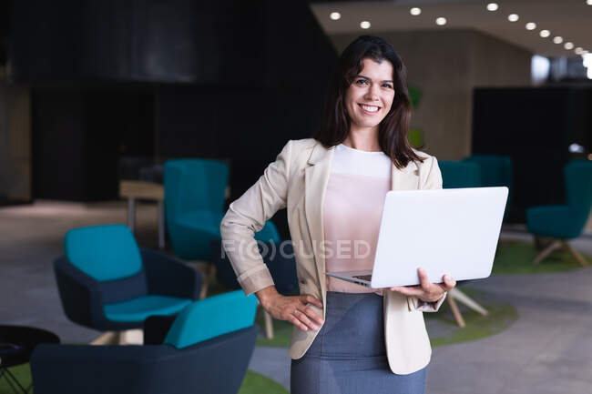 Portrait d'une femme d'affaires caucasienne tenant un ordinateur portable souriant tout en se tenant debout au bureau moderne. concept d'entreprise et de bureau — Photo de stock