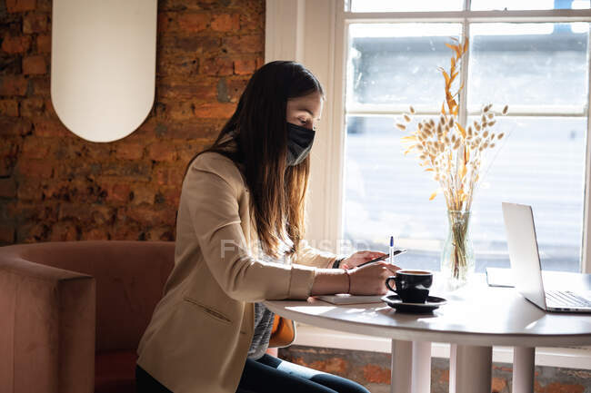 Cliente femenino caucásico con máscara facial sentada en la mesa, tomando notas. pequeño negocio de café independiente durante coronavirus covid 19 pandemia. - foto de stock