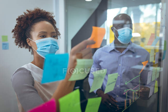 Two diverse business colleagues wearing face masks and taking notes on glass board. work at a modern office during covid 19 coronavirus pandemic. — Stock Photo