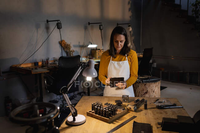 Сосредоточенная белая ювелирка в фартуке, стоящая за столом в мастерской, с помощью планшета. независимый ремесленный бизнес. — стоковое фото