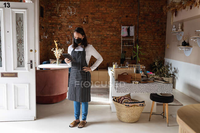 Кавказька жінка-власник бізнесу, одягнена в маску обличчя і фартух, за допомогою таблетки. Невеличкий незалежний кафе під час коронавірусної ковини 19. — стокове фото