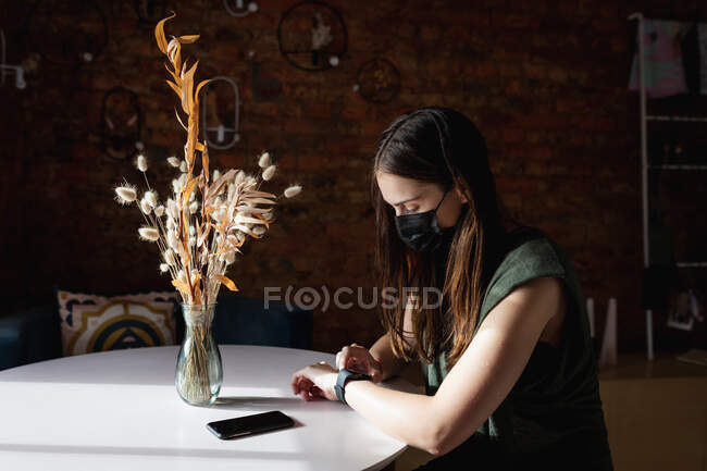 Белая клиентка в маске, сидит за столом, используя умные часы. Небольшой независимый бизнес кафе во время пандемии коронавируса. — стоковое фото