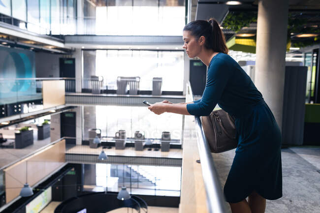 Mujer de negocios caucásica usando un teléfono inteligente mientras está de pie en el pasillo de la oficina moderna. concepto de negocio y oficina - foto de stock
