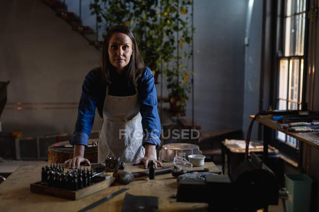 Кавказька жінка-ювелір стоїть за столом у майстерні, дивлячись на камеру. Незалежний ремісничий бізнес. — стокове фото