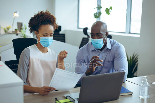Two diverse business colleagues wearing face masks and discussing. work at a modern office during covid 19 coronavirus pandemic. — Stock Photo