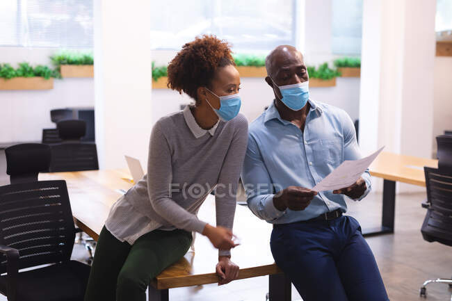 Dos diversos colegas de negocios masculinos y femeninos que usan máscaras faciales y tienen documentos. trabajar en una oficina moderna durante la pandemia de coronavirus covid 19. - foto de stock