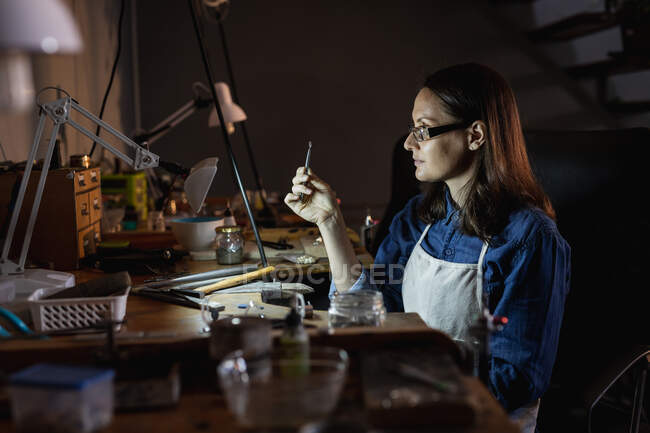 Jeweller feminino caucasiano vestindo avental, e óculos, sentado na mesa, fazendo jóias na oficina. negócios artesanais independentes. — Fotografia de Stock