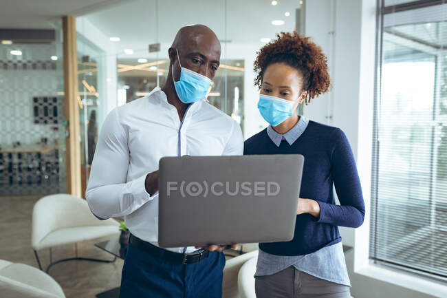 Dos diversos colegas de negocios con máscaras faciales y el uso de ordenador portátil. trabajar en una oficina moderna durante la pandemia de coronavirus covid 19. - foto de stock