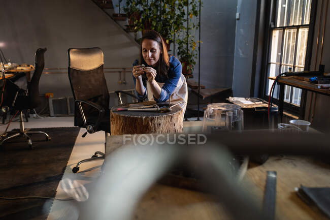 Gioielleria femminile caucasica sorridente appoggiata sul tavolo, che controlla i gioielli in officina. artigianato artigianale indipendente. — Foto stock