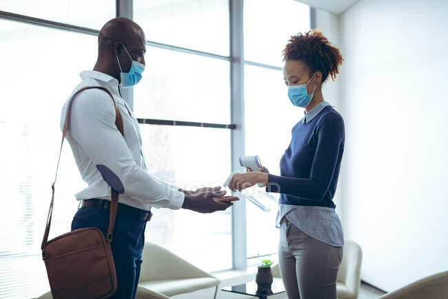 Dos colegas de negocios diversos que usan máscaras faciales que desinfectan las manos y toman temperatura. trabajar en una oficina moderna durante la pandemia de coronavirus covid 19. - foto de stock