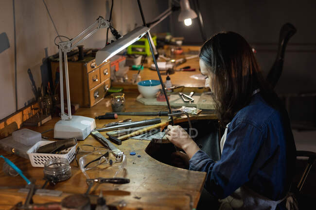 Kaukasische Juwelierin sitzt am Schreibtisch, hält Schmuckwerkzeuge in der Hand, fertigt Schmuck in der Werkstatt. Selbstständiger Handwerksbetrieb. — Stockfoto