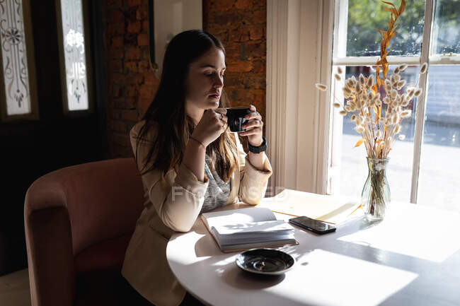 Кавказька жінка-клієнтка сидить за столом біля вікна і п'є каву. маленьке незалежне кафе.. — стокове фото