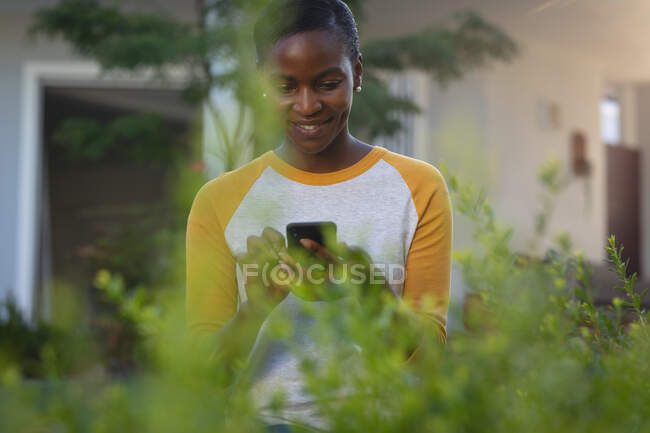 Sorridente donna afroamericana in piedi in giardino soleggiato utilizzando lo smartphone. trascorrere del tempo libero a casa. — Foto stock