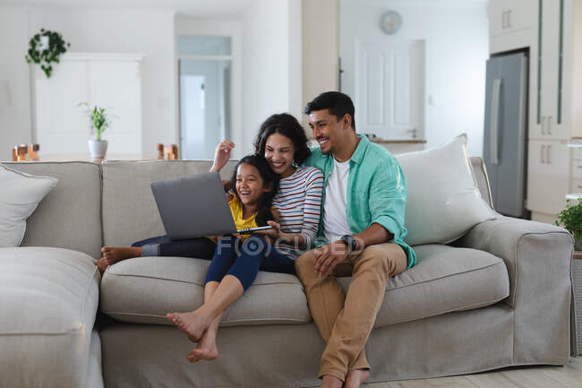 Улыбающиеся латиноамериканская мать, отец и дочь сидят на диване, используя ноутбук вместе. Семья проводит время вместе дома. — стоковое фото