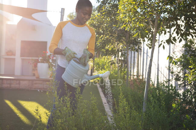 Улыбающаяся африканская американка, садоводческая женщина, стоящая в солнечном саду, поливая растения лейкой. проводить свободное время дома. — стоковое фото