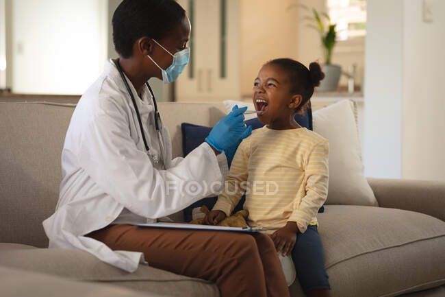 African american female doctor in face mask giving girl patient covid swab test at home. medical and healthcare services during coronavirus covid 19 pandemic. — Stock Photo