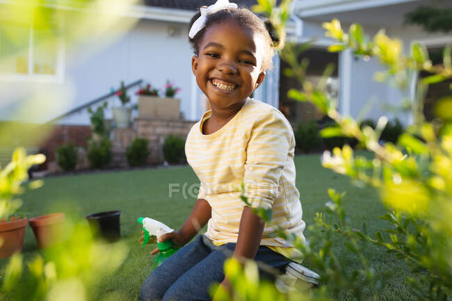 Porträt eines lächelnden afrikanisch-amerikanischen Mädchens beim Gärtnern, kniend, mit Wasserspray in der Hand im sonnigen Garten. Freizeit zu Hause verbringen. — Stockfoto