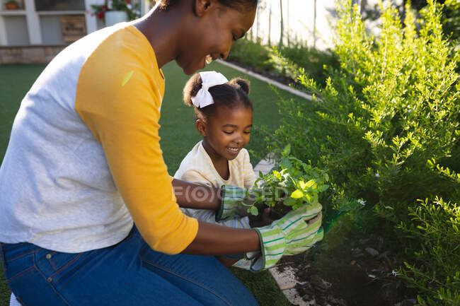 Счастливая африканская американская мать и дочь стоят на коленях, ухаживая за растениями в солнечном саду. Семья проводит время вместе дома. — стоковое фото