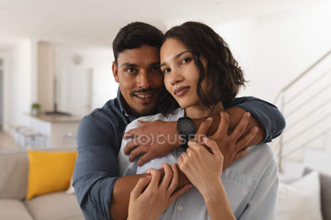 Retrato de feliz pareja hispana abrazándose en la sala de estar, mirando a la cámara. en casa en aislamiento durante el bloqueo de cuarentena. - foto de stock