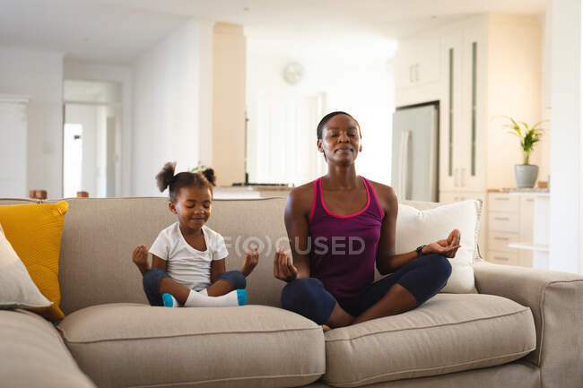 Feliz madre afroamericana y su hija practicando yoga, meditando en el sofá en la sala de estar. familia pasar tiempo juntos en casa. - foto de stock