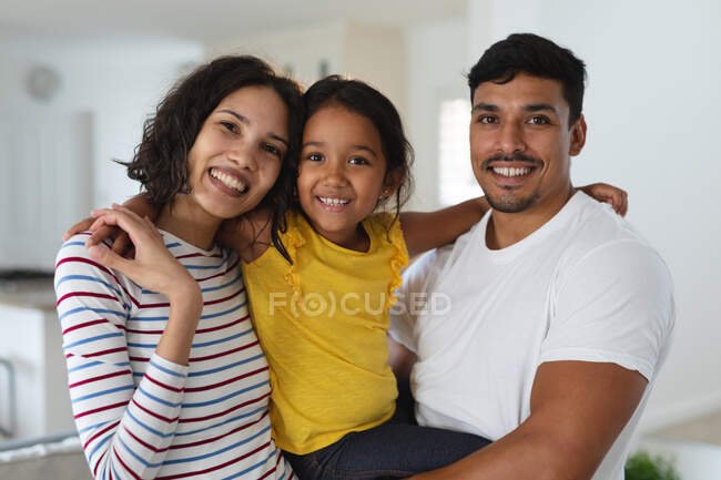 Portrait of smiling hispanic father embracing with wife and daughter standing in living room. happy family at home together. — Stock Photo