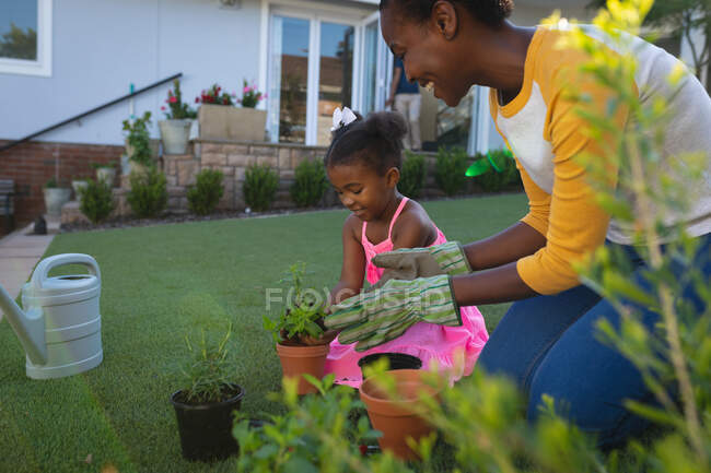 Heureuse mère et fille afro-américaine agenouillée tendant à des plantes en pot dans le jardin. passer du temps ensemble à la maison. — Photo de stock