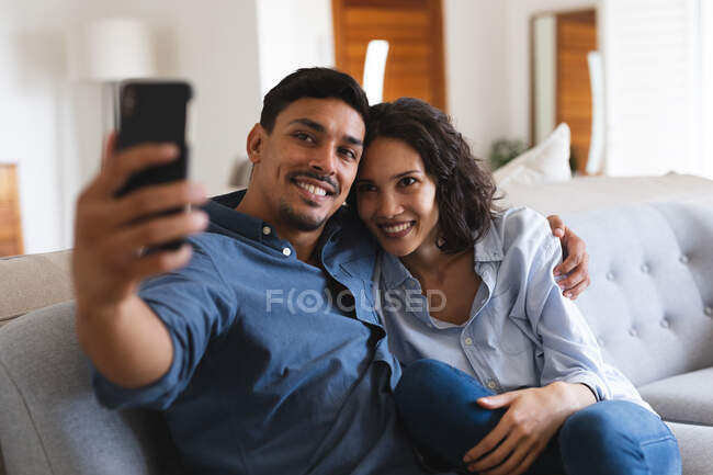 Glückliches hispanisches Paar, das auf der Couch im Wohnzimmer sitzt, Selfie macht und lächelt. Zeit gemeinsam zu Hause verbringen. — Stockfoto