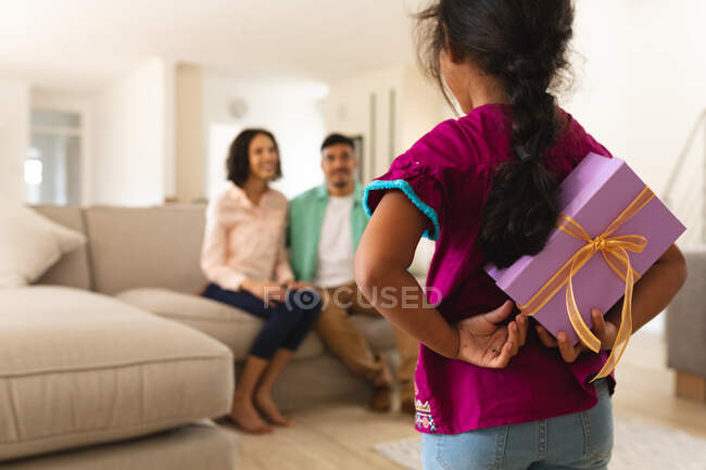 Hispanic parents sitting on couch with daughter standing in foreground hiding present behind back. spending time together at home. — Stock Photo