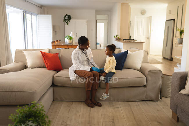 Sorrindo afro-americana médica visitando paciente menina em casa, sentado no sofá falando. serviços médicos e de saúde, visita domiciliária médica. — Fotografia de Stock