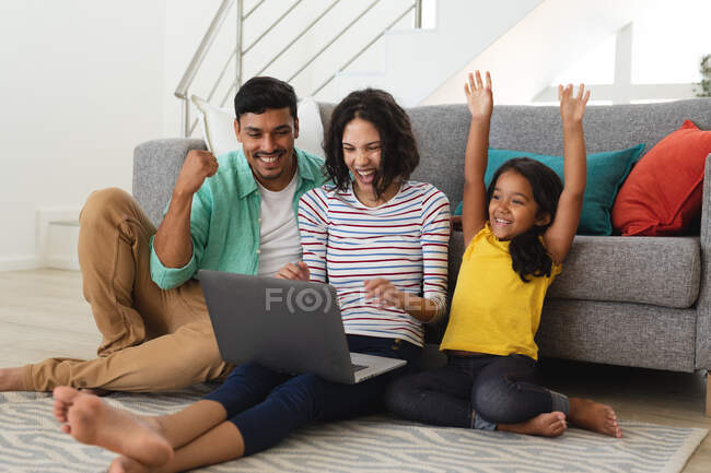 Animando a la madre hispana, padre e hija sentados en el piso de la sala de estar usando un portátil juntos. familia pasar tiempo juntos en casa. - foto de stock