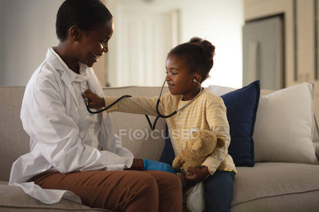 Усміхнена афроамериканська жінка-лікар, яка відвідує пацієнта вдома, грає зі стетоскопом. медичні та медичні послуги, візит лікаря додому . — стокове фото