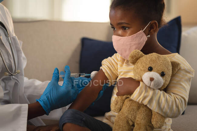 African american female doctor giving covid vaccination to girl patient in face mask at home. medical and healthcare services during coronavirus covid 19 pandemic. — Stock Photo