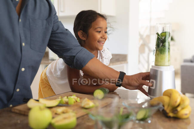 Lächelnde hispanische Tochter stützt sich auf den Tresen und beobachtet den Vater bei der Zubereitung eines Gesundheitsgetränks in der Küche. Zuhause in Isolation während der Quarantäne. — Stockfoto